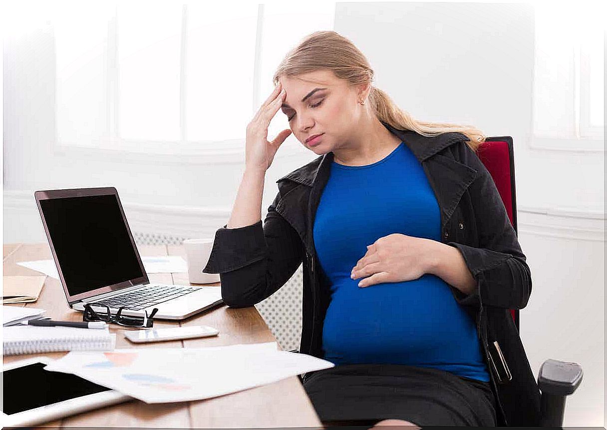 A pregnant woman sitting at her workplace holding her forehead.